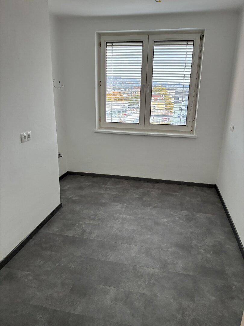 Leeres Zimmer mit dunklem Bodenbelag und Fenster, das eine städtische Aussicht ermöglicht.