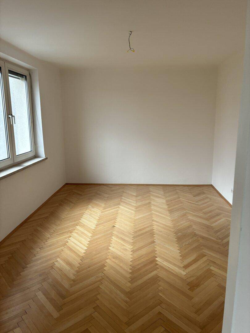 Geräumiges, helles Zimmer mit klassischem Fischgrätparkett und einem großen Fenster.
