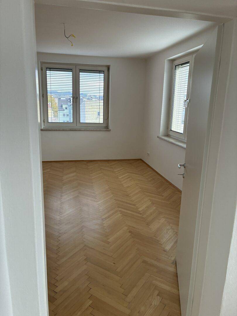 Helles Zimmer mit Fischgrätparkett und zwei Fenstern, die viel Tageslicht hereinlassen.