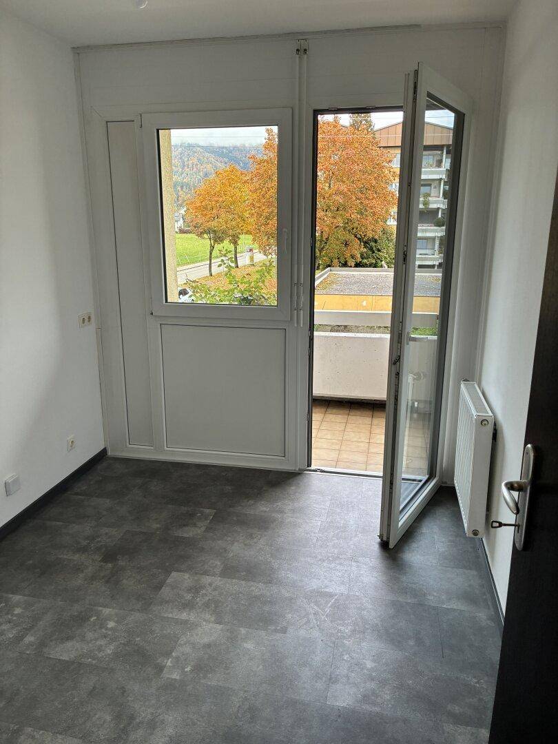Helles Zimmer mit Balkonzugang, dunklem Fliesenboden und Blick ins Grüne.