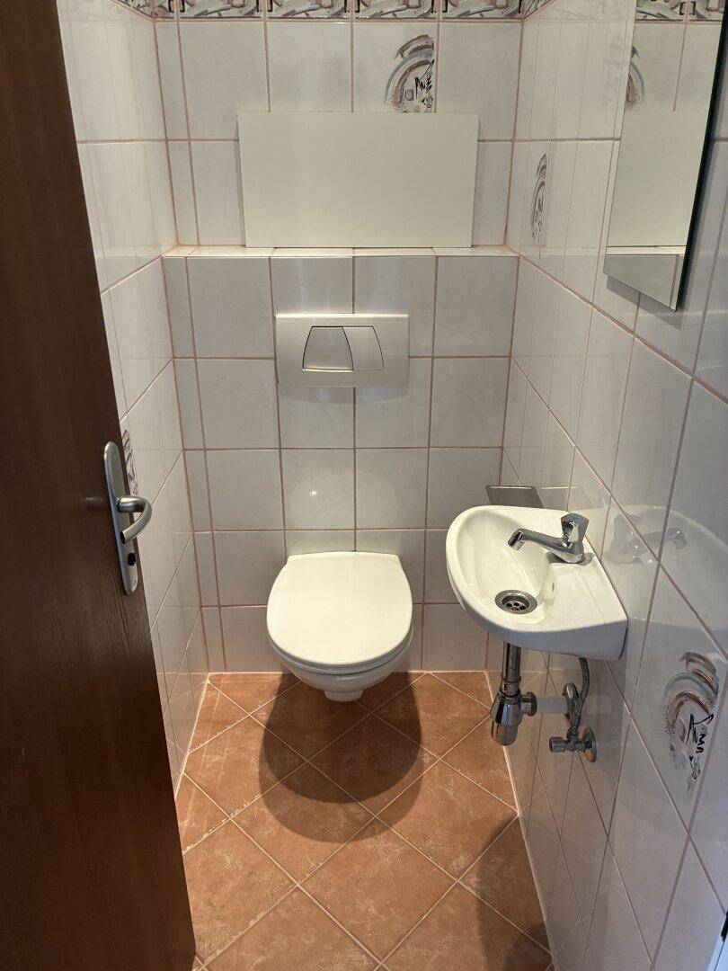 Gäste-WC mit Wand-WC, kleinem Waschbecken und hellen Fliesen an Wänden und Boden.