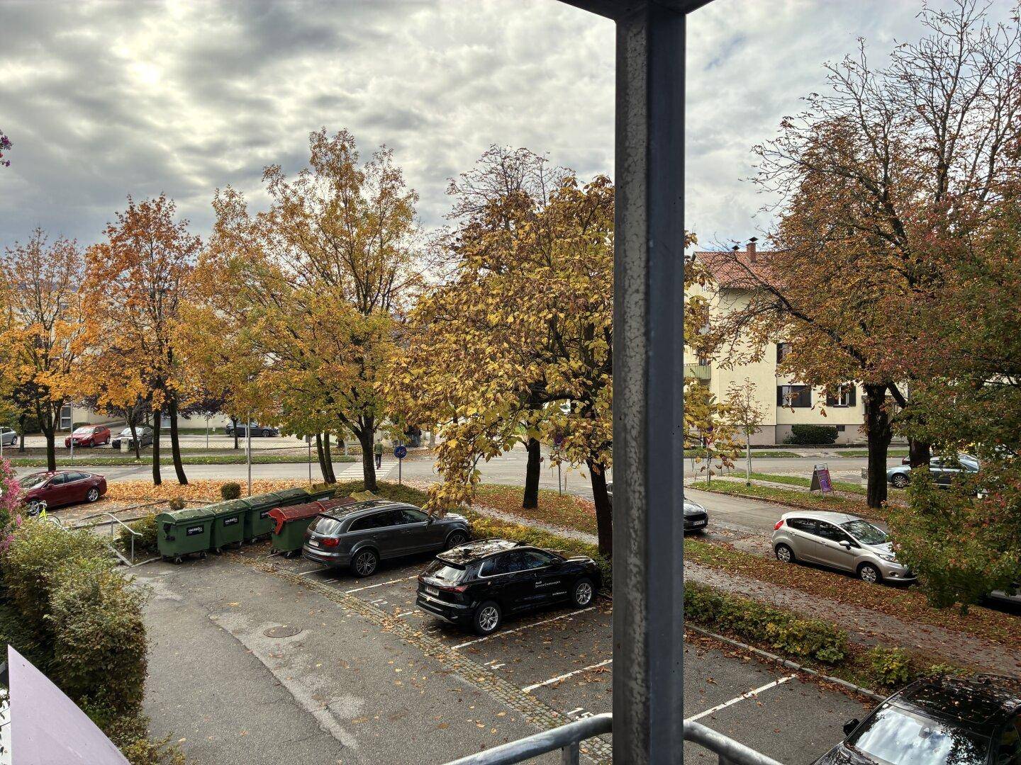 Blick vom Balkon auf einen Parkplatz und herbstliche Bäume an einem bewölkten Tag.