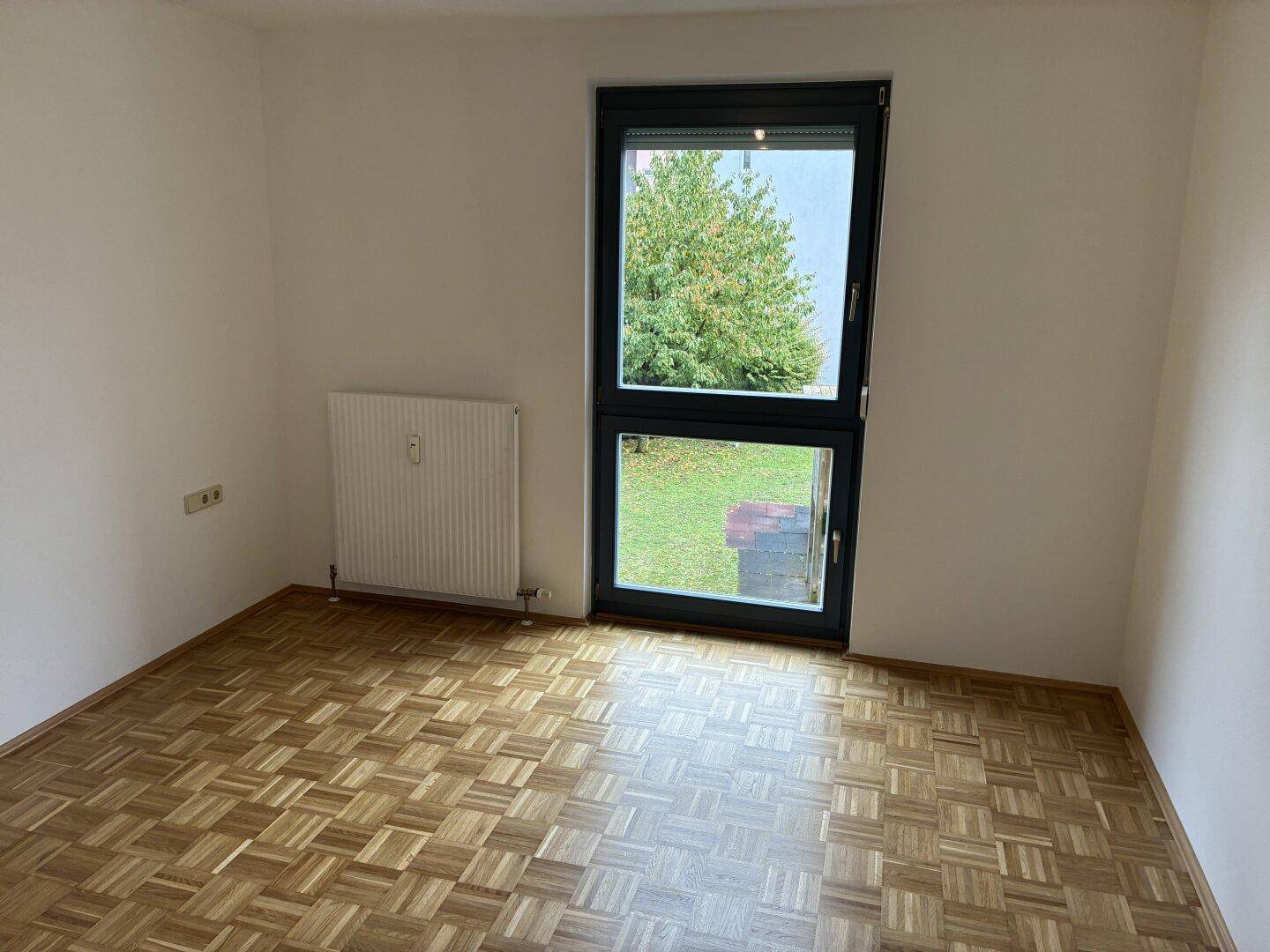 Helles Schlafzimmer mit Parkettboden, Heizkörper und Blick ins Grüne.