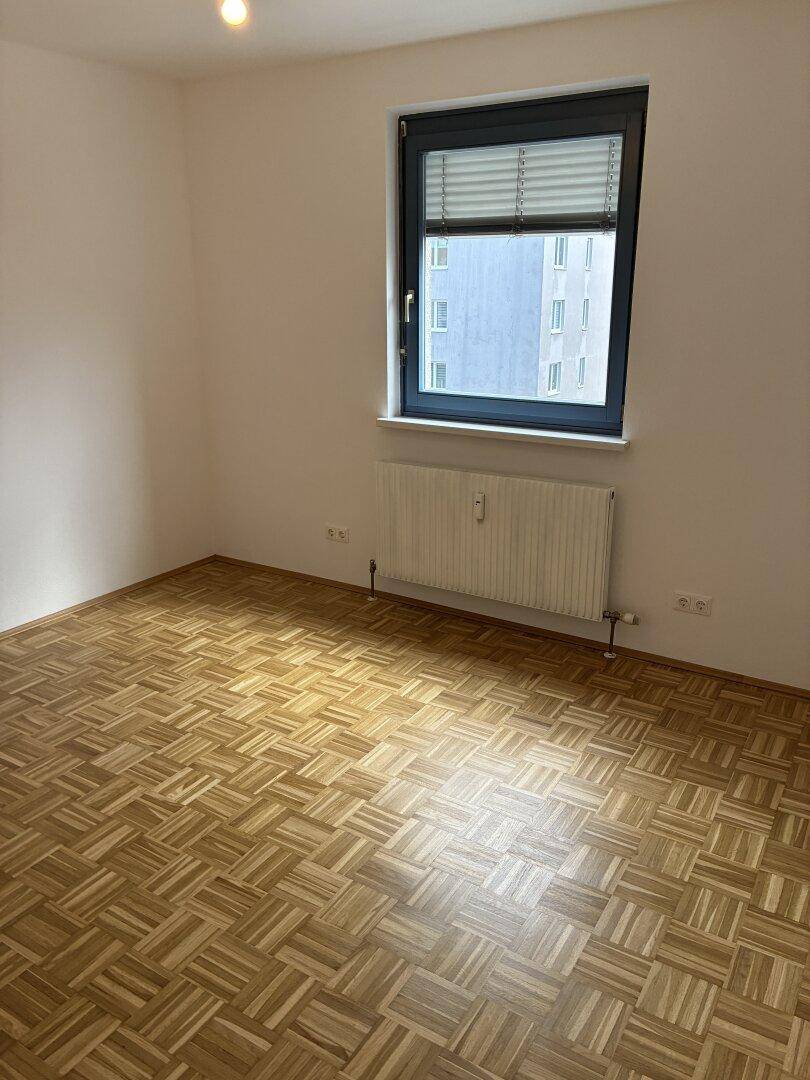 Leeres Zimmer mit Parkettboden und einem Fenster, das Tageslicht hereinlässt.
