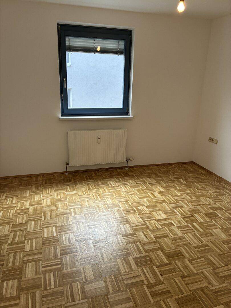 Helles, leeres Zimmer mit Parkettboden und einem großen Fenster für viel Tageslicht.