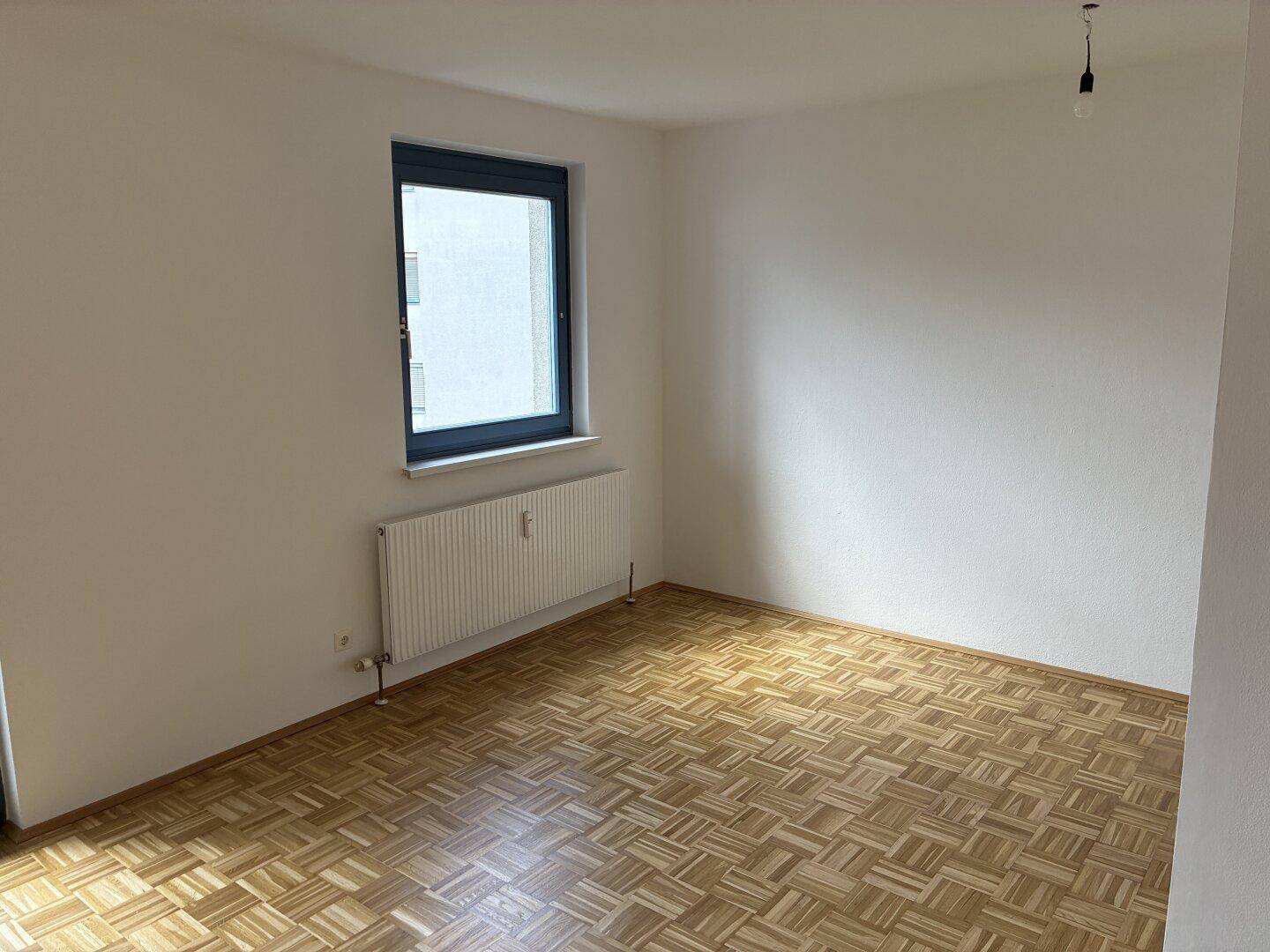 Leeres Zimmer mit Parkettboden und einem Fenster, das eine helle Atmosphäre schafft.