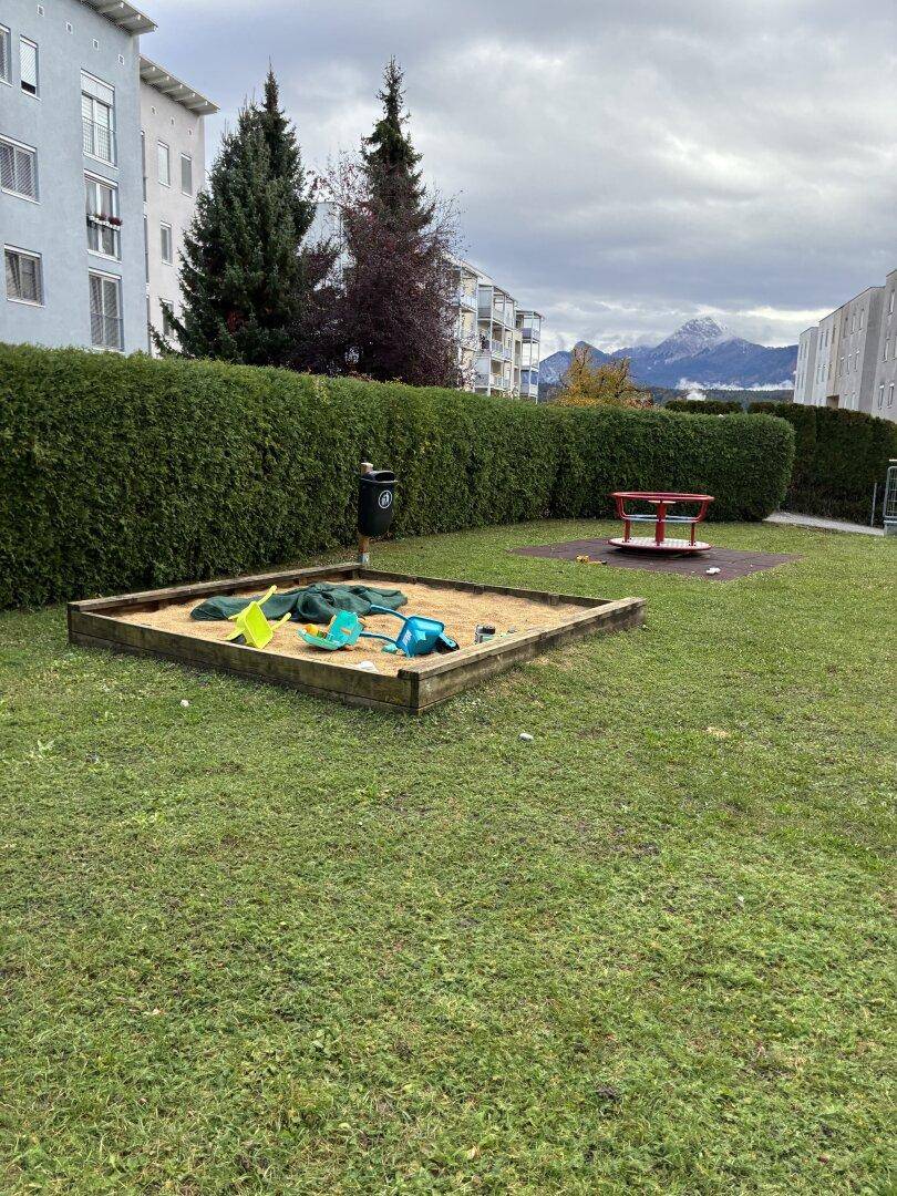 Grüner Außenbereich mit Sandkasten und Spielgeräten, umgeben von einer Hecke.