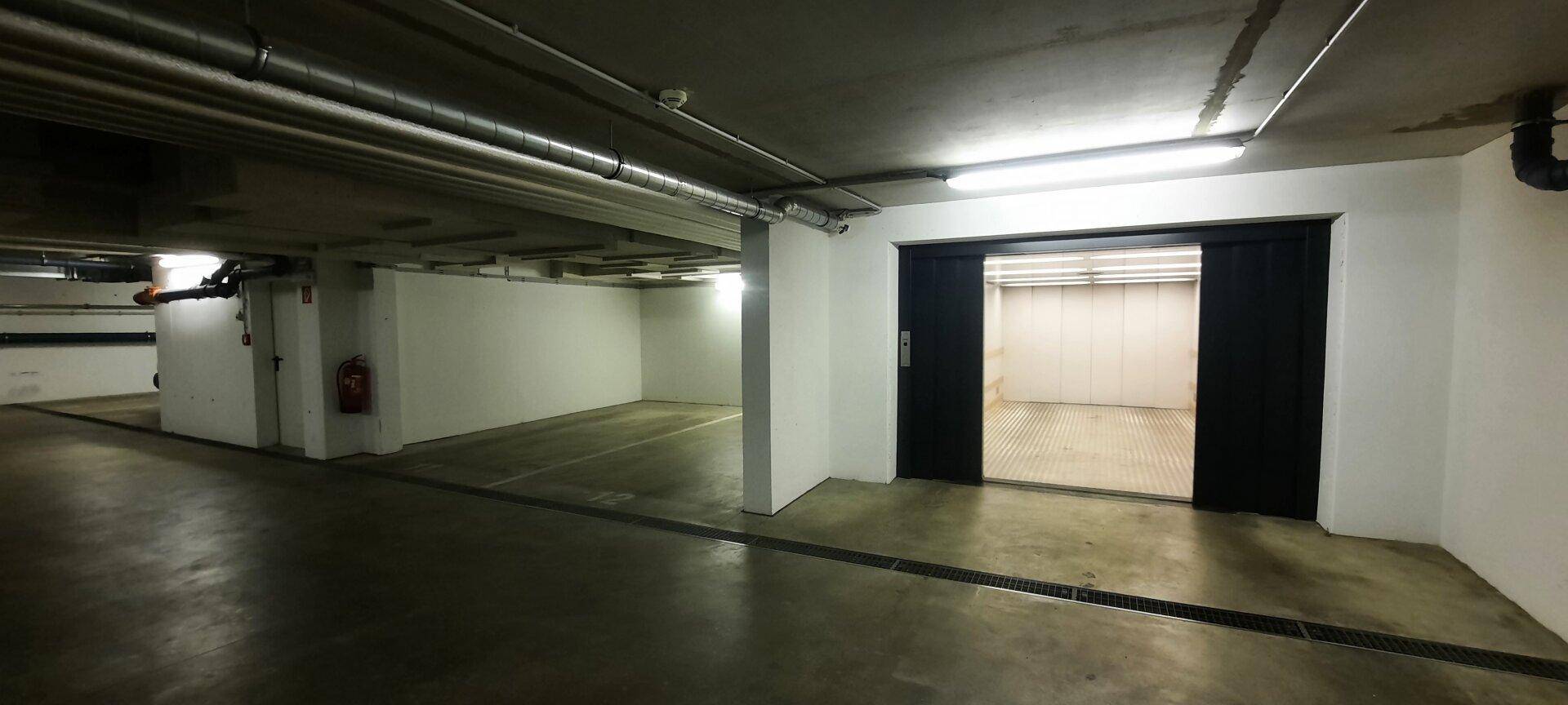 Weitläufige Tiefgarage mit Betonboden und offenem Zugang zum Autoaufzug, beleuchtet von Neonröhren.