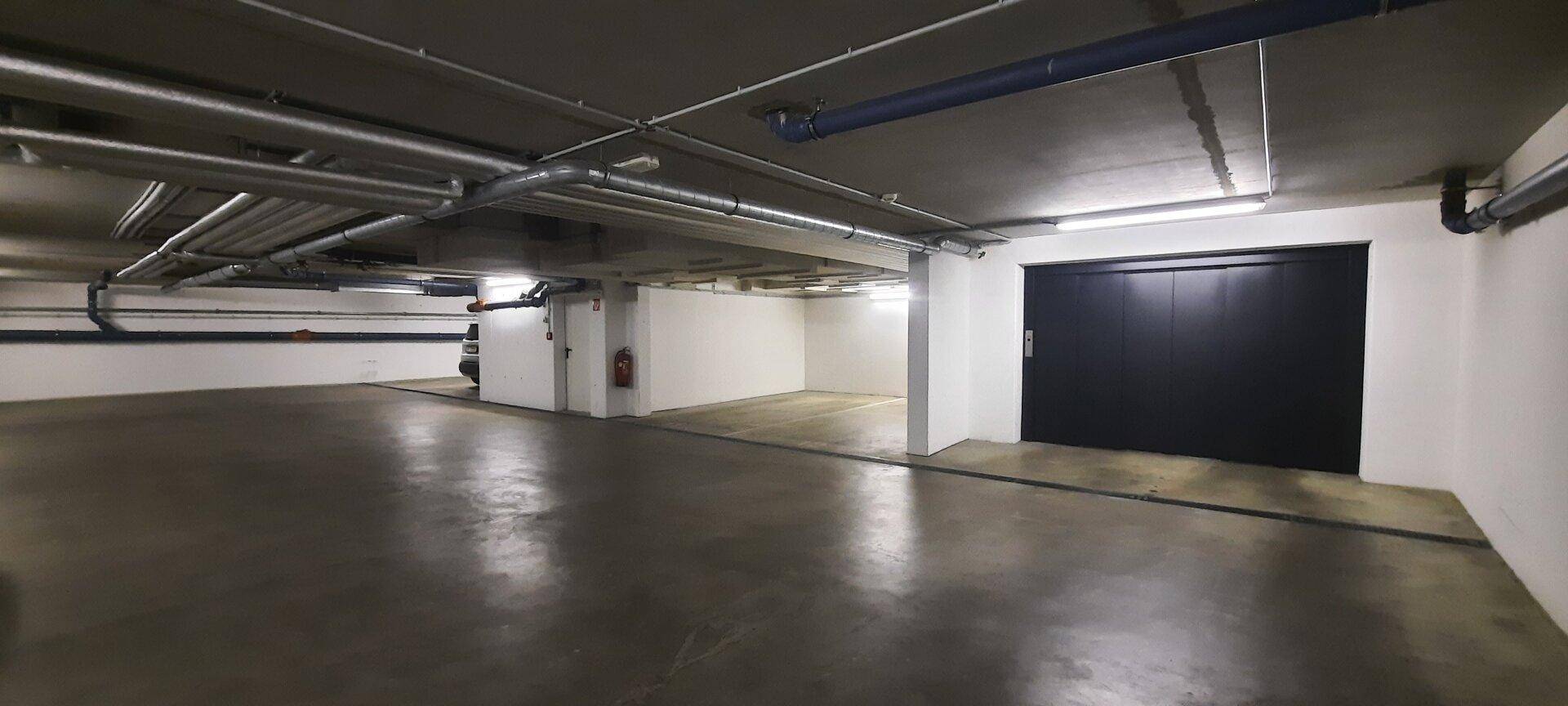 Geräumige Tiefgarage mit geschlossener Autoaufzugtür und sichtbaren Installationen an der Decke.