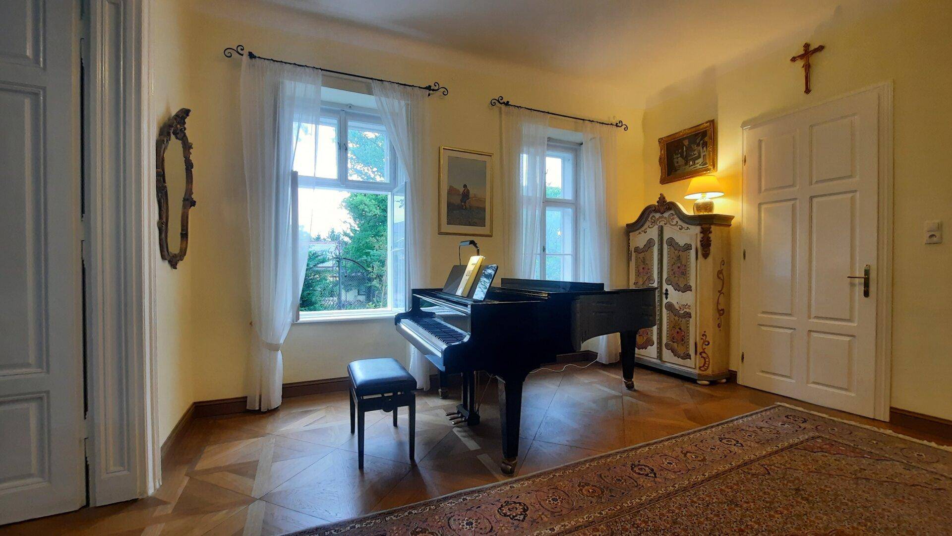 Musikzimmer mit Flügel, Parkettboden und hohen Fenstern, die viel Licht hereinlassen.