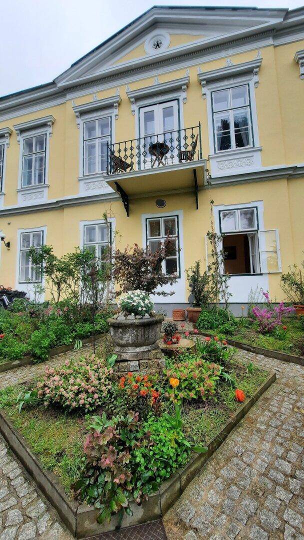 Gepflegter Vorgarten mit Blumenbeeten, einem Brunnen und gepflasterten Wegen vor dem Gebäude.