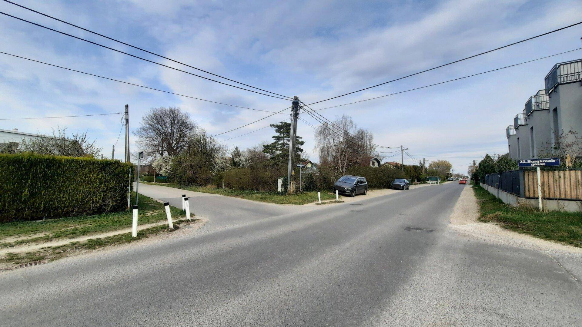Weitläufige Straßenkreuzung in ländlicher Umgebung mit Hecke und modernen Gebäuden im Hintergrund.