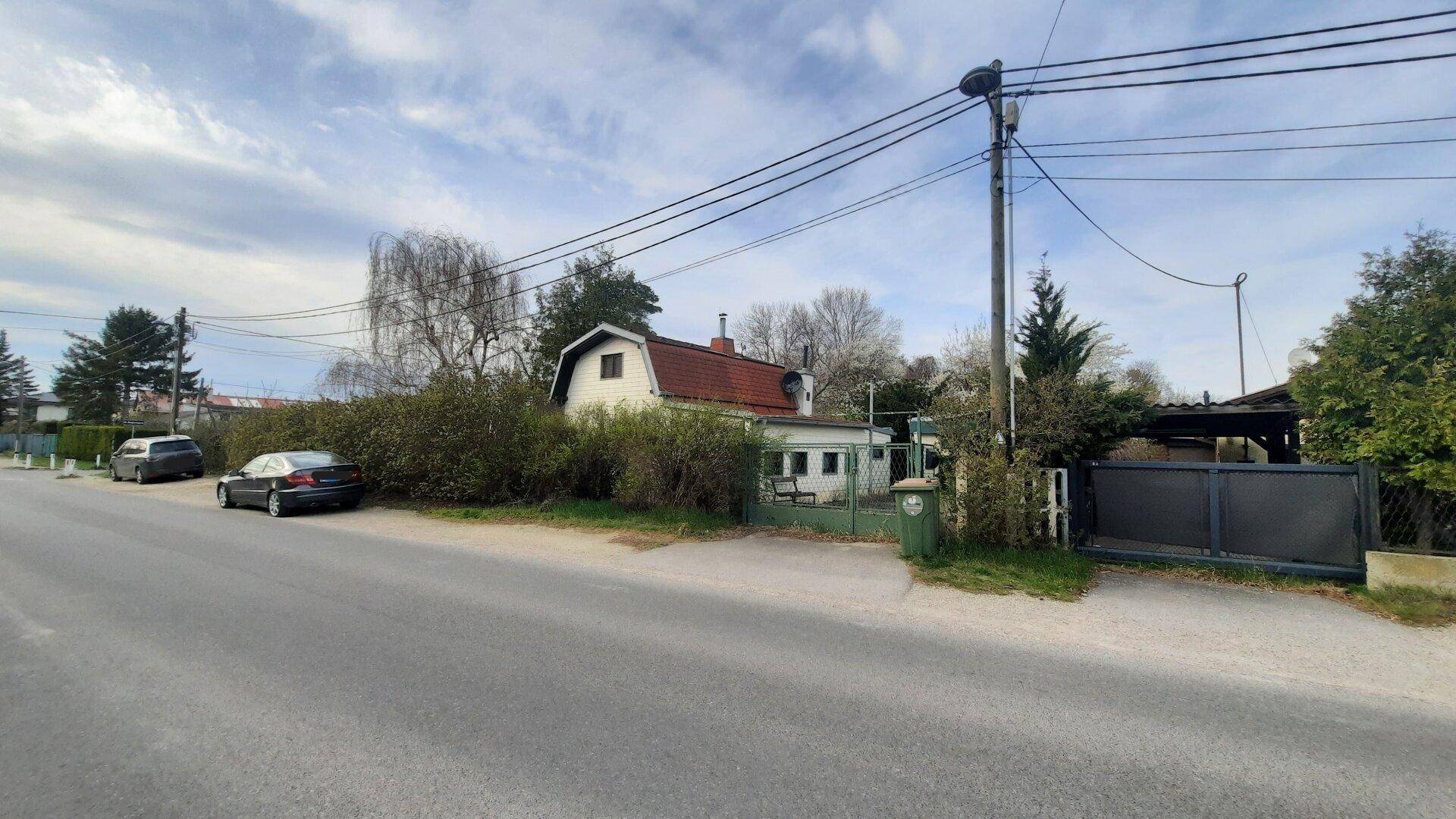 Ländliches Haus mit rotem Ziegeldach, umgeben von Bäumen und Büschen, an einer Straße gelegen.