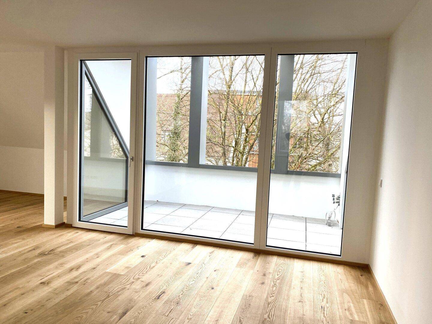 Heller Raum mit Holzboden und breiter Fensterfront, die auf eine geflieste Terrasse blickt.