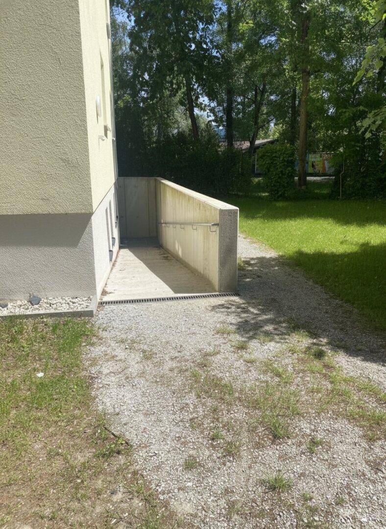 Außenbereich mit einer Rampe, die zu einem Kellereingang führt, umgeben von Grünflächen.