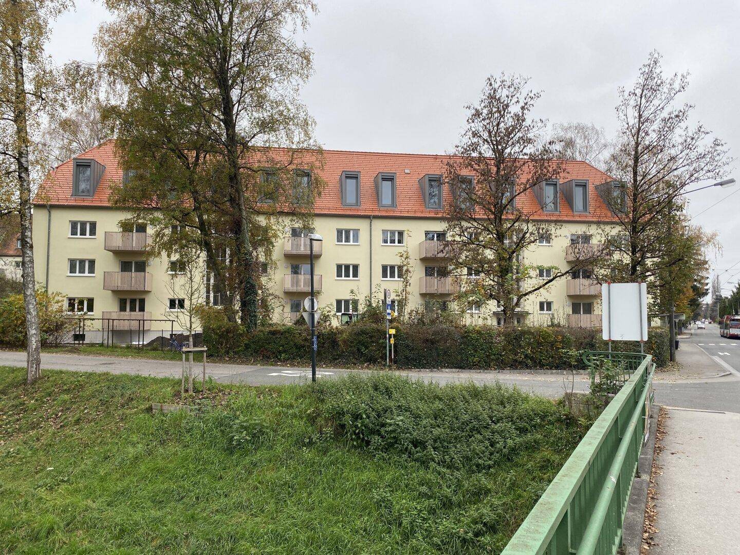 Mehrfamilienhaus mit Balkonen, umgeben von Bäumen, gesehen von der anderen Straßenseite mit Brücke.
