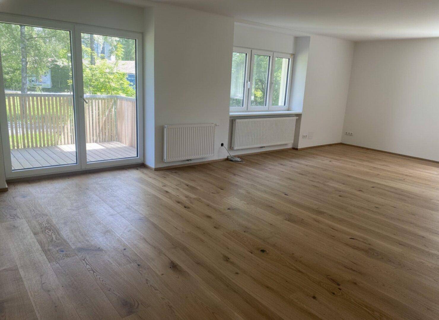 Geräumiger, leerer Raum mit Holzboden, großen Fenstern und Zugang zu einem Außenbereich.