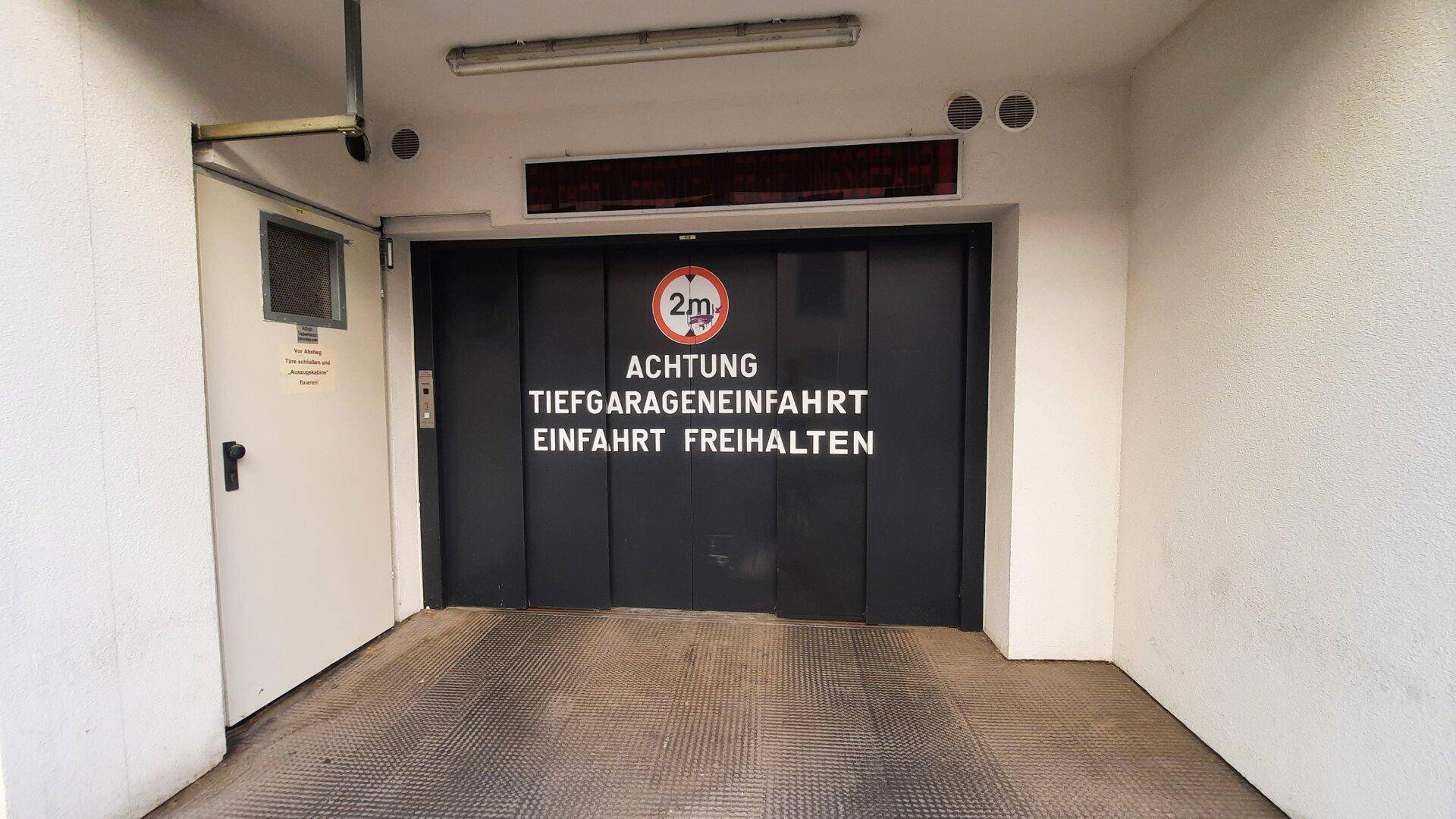 Eingang zur Tiefgarage mit schwarzem Tor und Hinweisschildern, darunter eine Höhenbegrenzung von 2m.