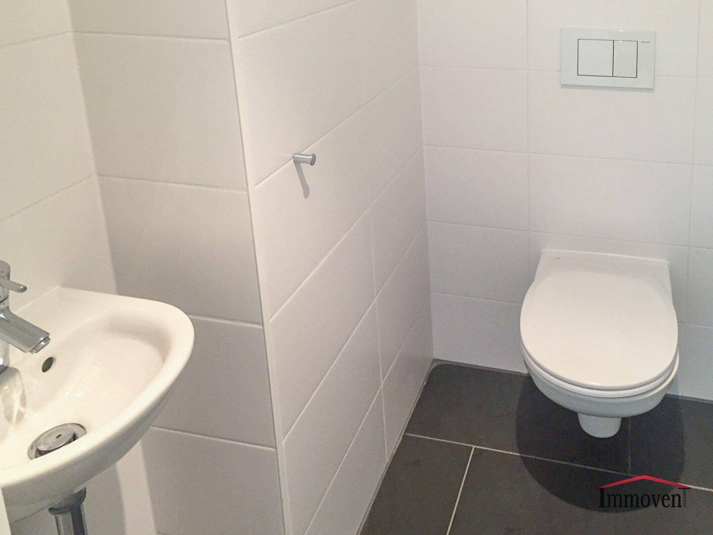 Separates WC mit wandhängender Toilette, kleinem Handwaschbecken und dunklen Bodenfliesen.