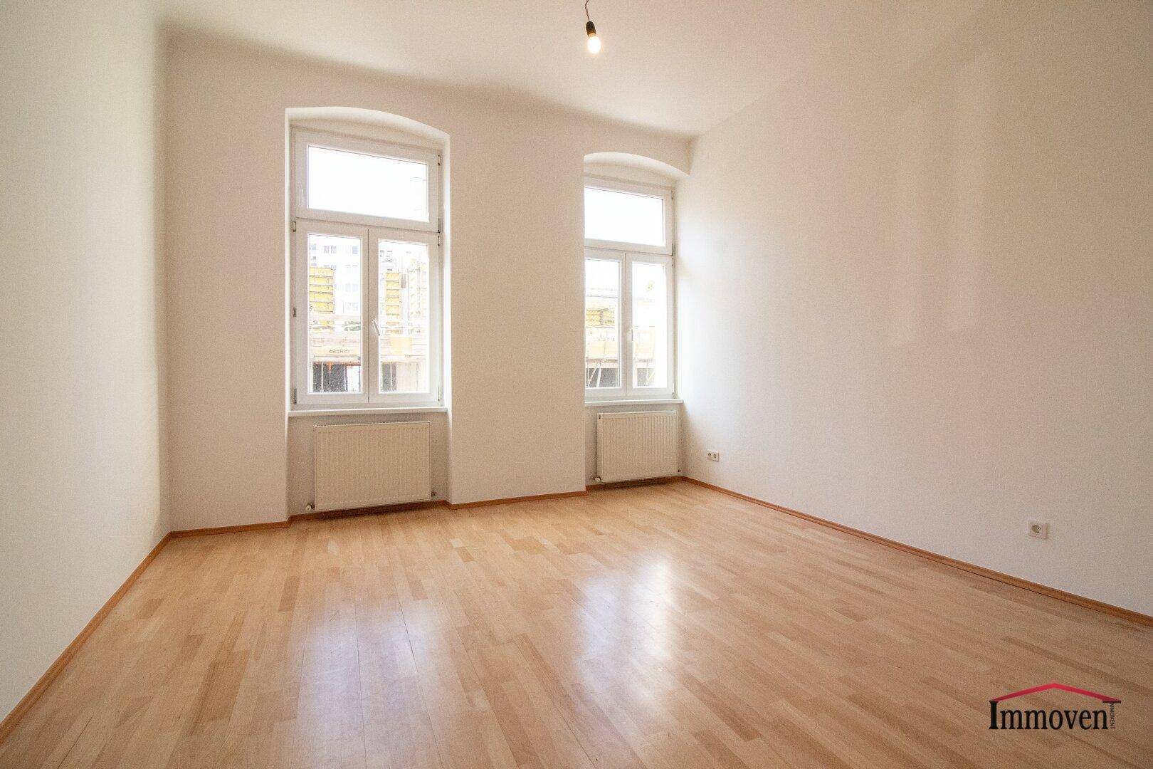 Leerer Raum mit hellem Parkettboden und zwei hohen Fenstern, die für eine freundliche Atmosphäre sorgen.
