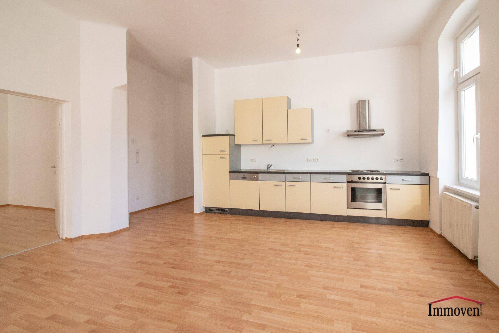 Offene Küche mit beigefarbenen Schränken, integrierten Geräten und hellem Parkettboden in einem geräumigen Zimmer.