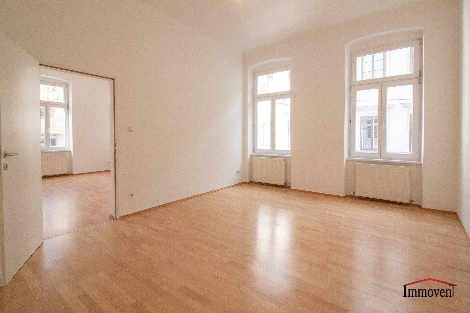 Heller Wohnraum mit Parkettboden und Zugang zu einem weiteren Zimmer, durch große Fenster gut belichtet.