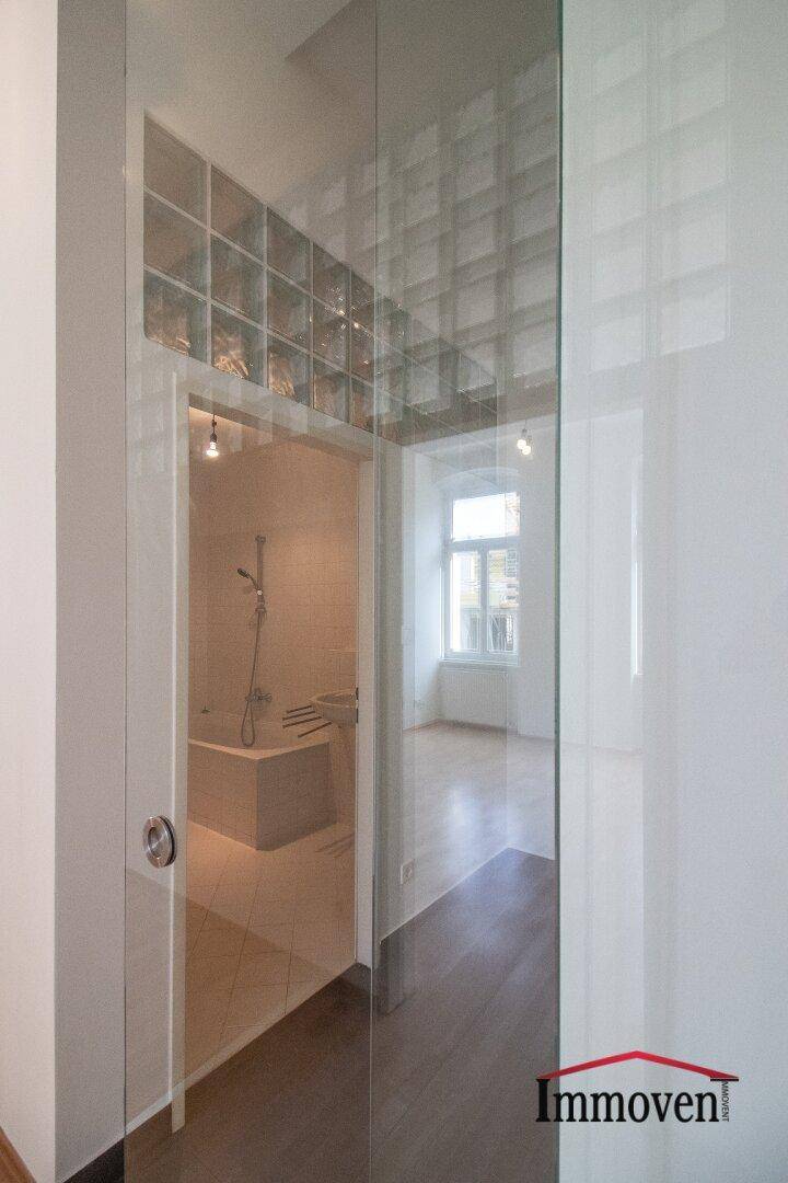 Blick durch eine Glasschiebetür in ein hell gefliestes Badezimmer mit Badewanne und Dusche.