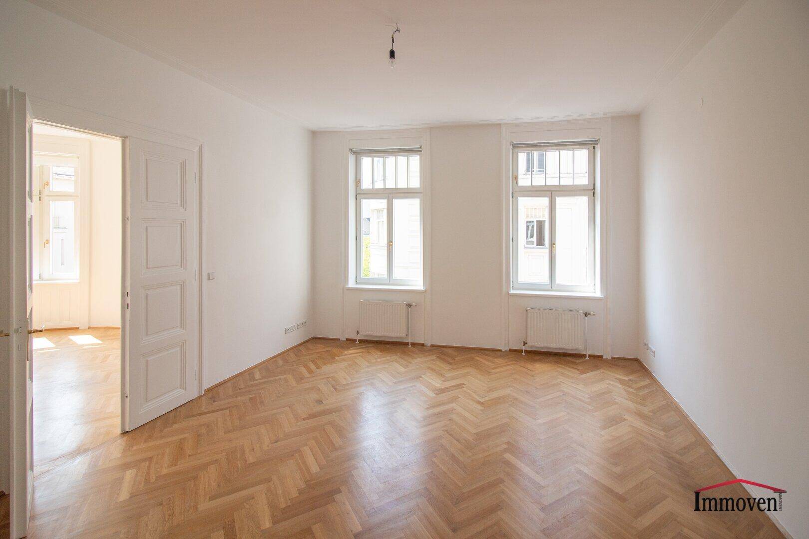 Geräumiger Wohnbereich mit Fischgrätparkett, weißen Wänden und einem Durchgang zu einem weiteren Zimmer.