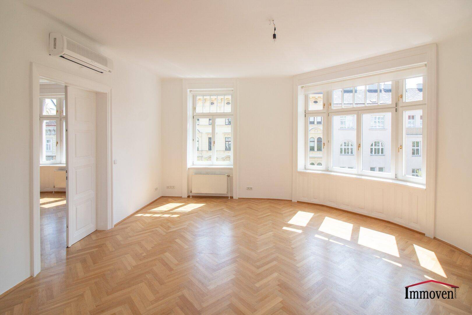 Geräumiger Wohnbereich mit Fischgrätparkett, weißen Wänden und einem Durchgang zu einem weiteren Zimmer.