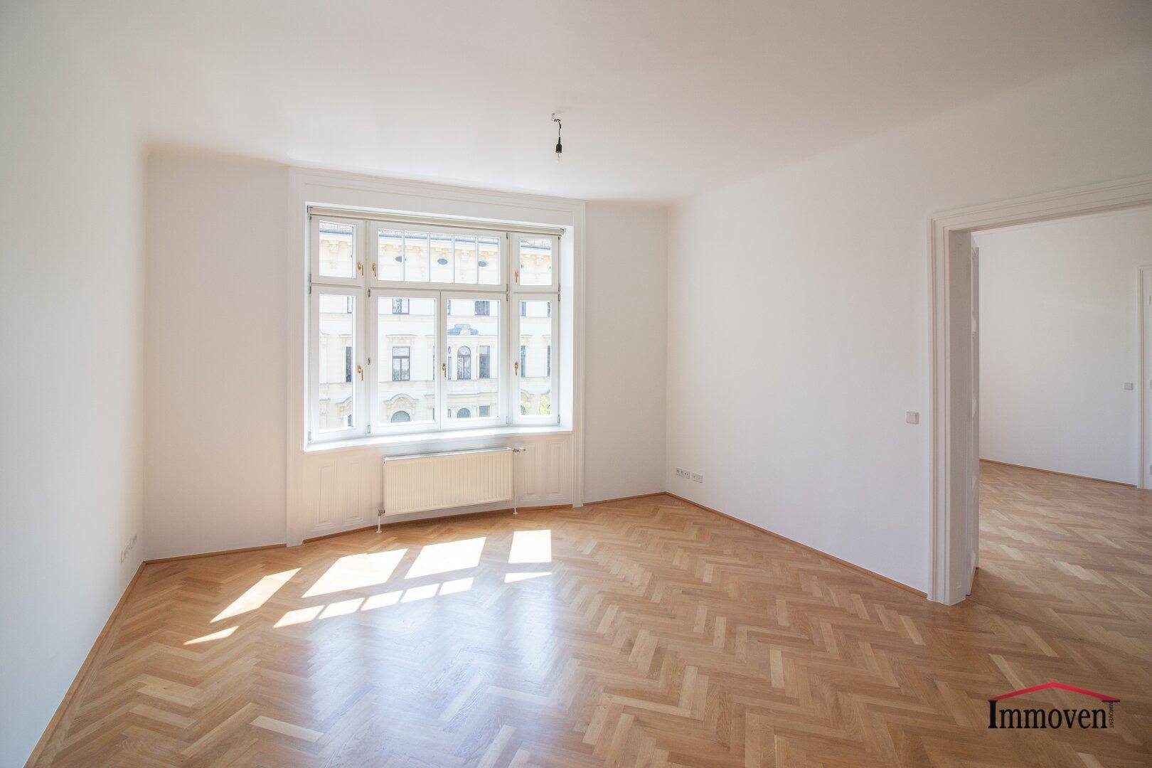 Lichtdurchfluteter Wohnraum mit klassischem Fischgrätparkett und einem breiten Fenster mit Blick nach draußen.