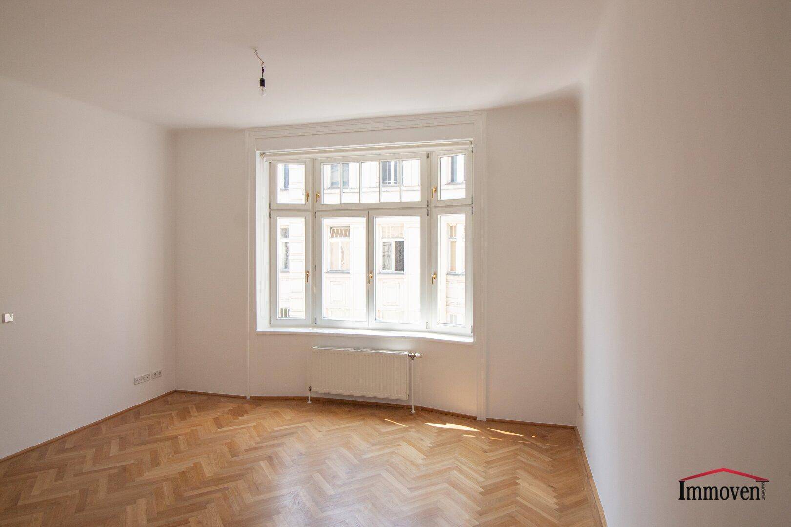 Geräumiger Wohnbereich mit Fischgrätparkett und einem großen Fenster, das den Raum erhellt.