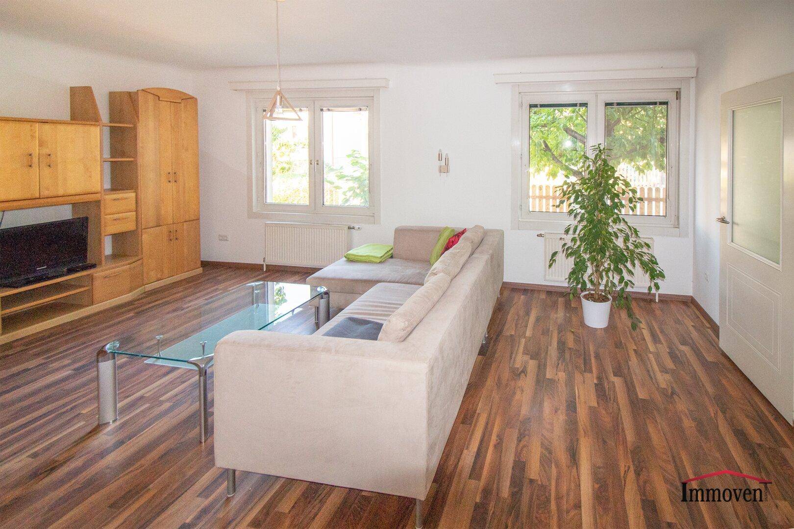 Geräumiges Wohnzimmer mit Laminatboden, beigem Sofa und großen Fenstern mit Grünblick.