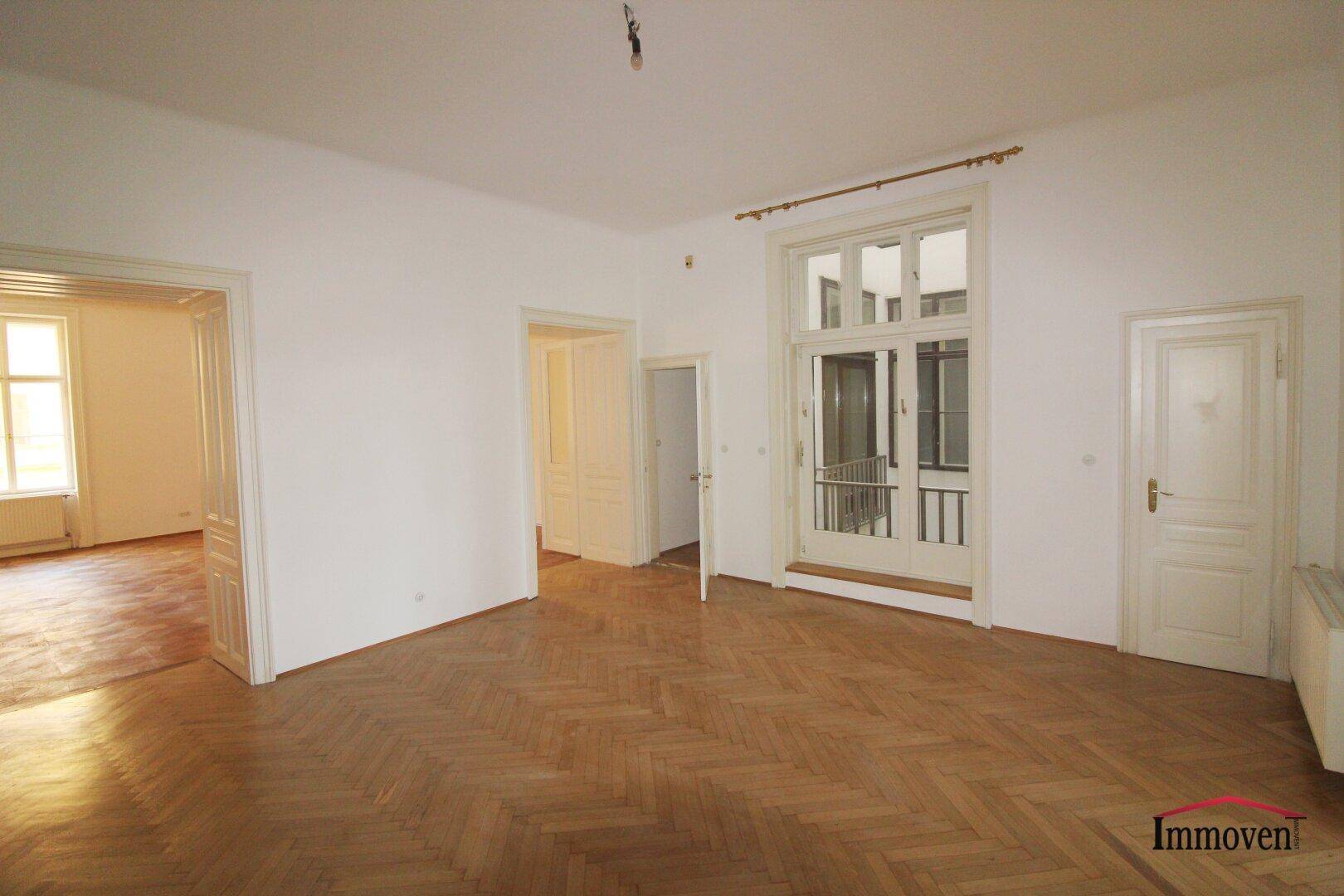 Geräumiger Raum mit Fischgrätparkett, hohen Decken und einer großen Fenstertür, die auf einen Balkon führt.