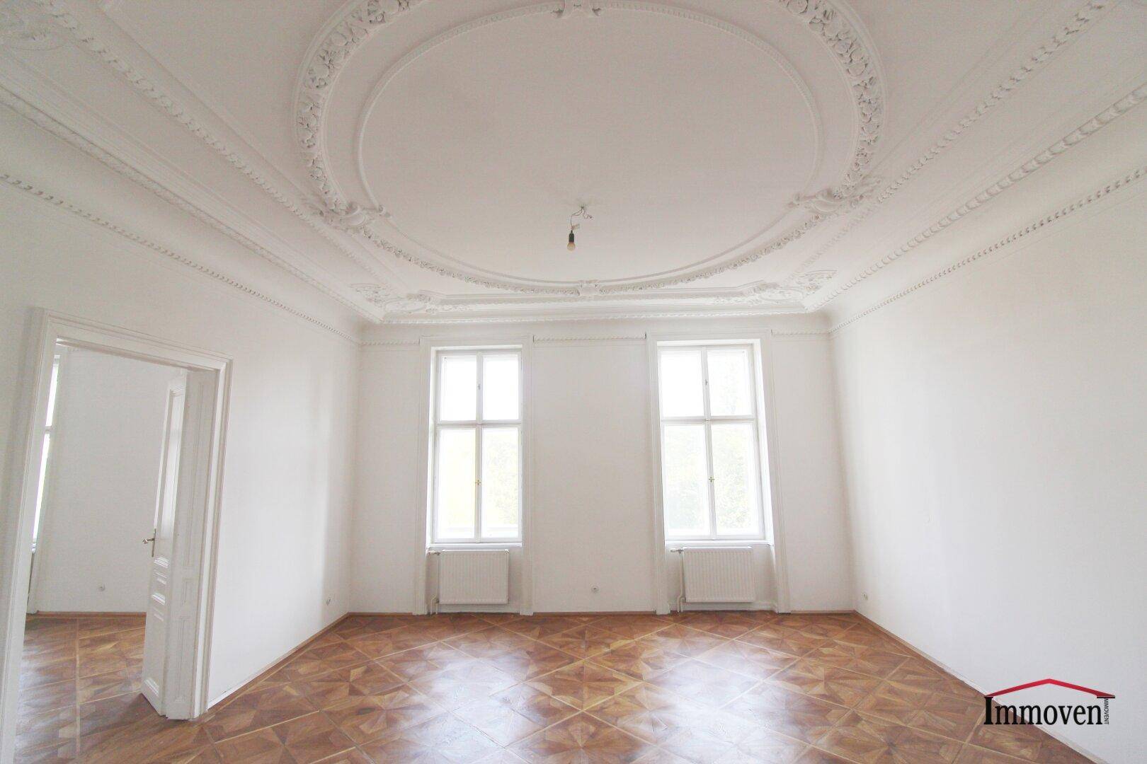 Großes, helles Zimmer mit klassischem Parkettboden und aufwendigem Deckenstuck. Zwei hohe Fenster lassen viel Licht herein.