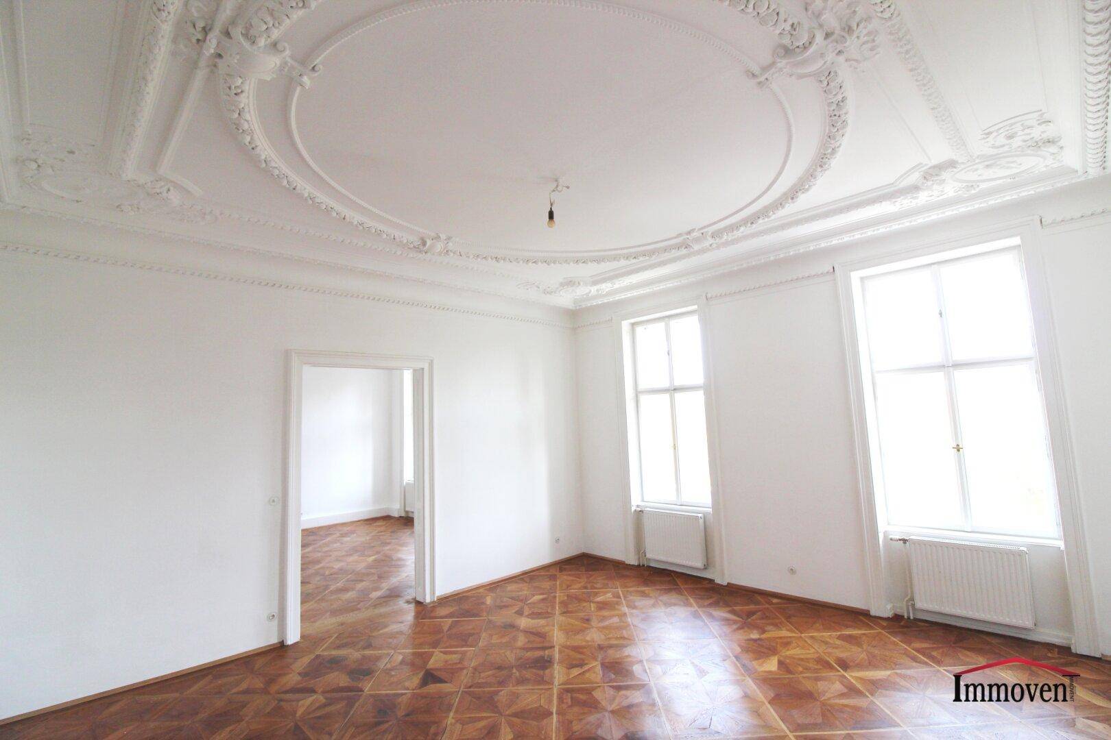 Helles Zimmer mit kunstvollem Deckenstuck, Fischgrätparkett und hohen Fenstern. Eine offene Tür führt in einen weiteren Raum.