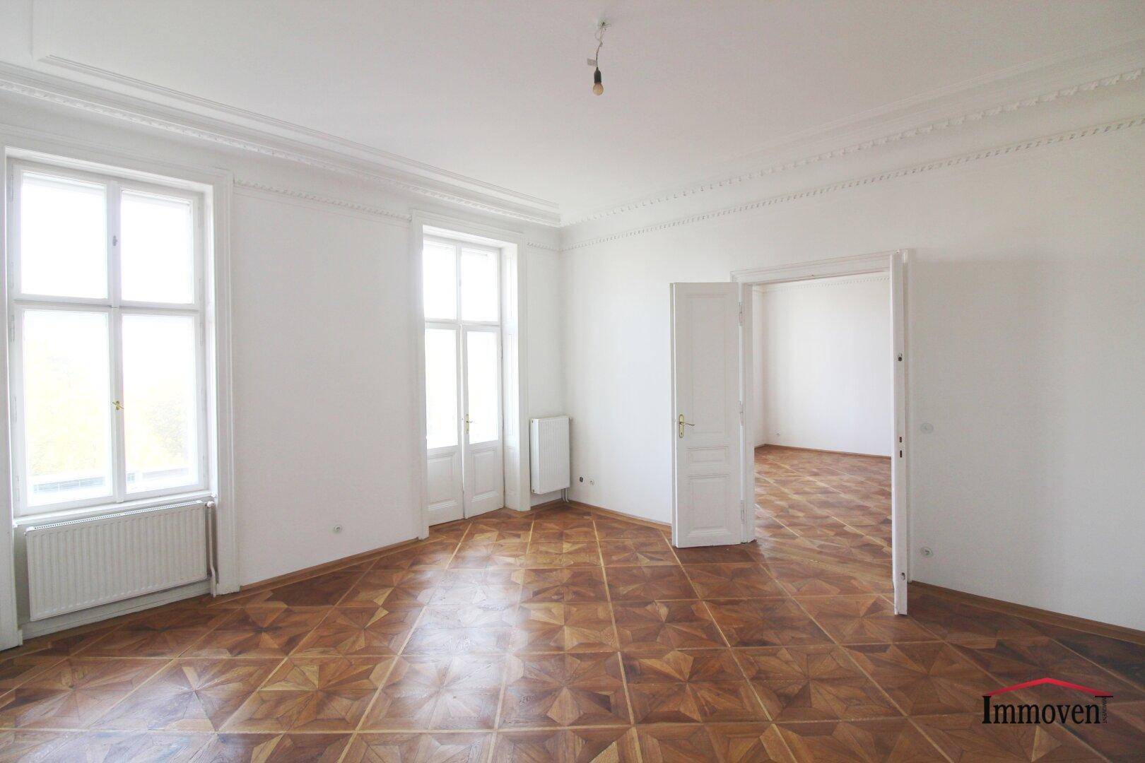 Geräumiges Zimmer mit elegantem Parkettboden, hohen Fenstern und einer Doppeltür zu einem weiteren Raum.