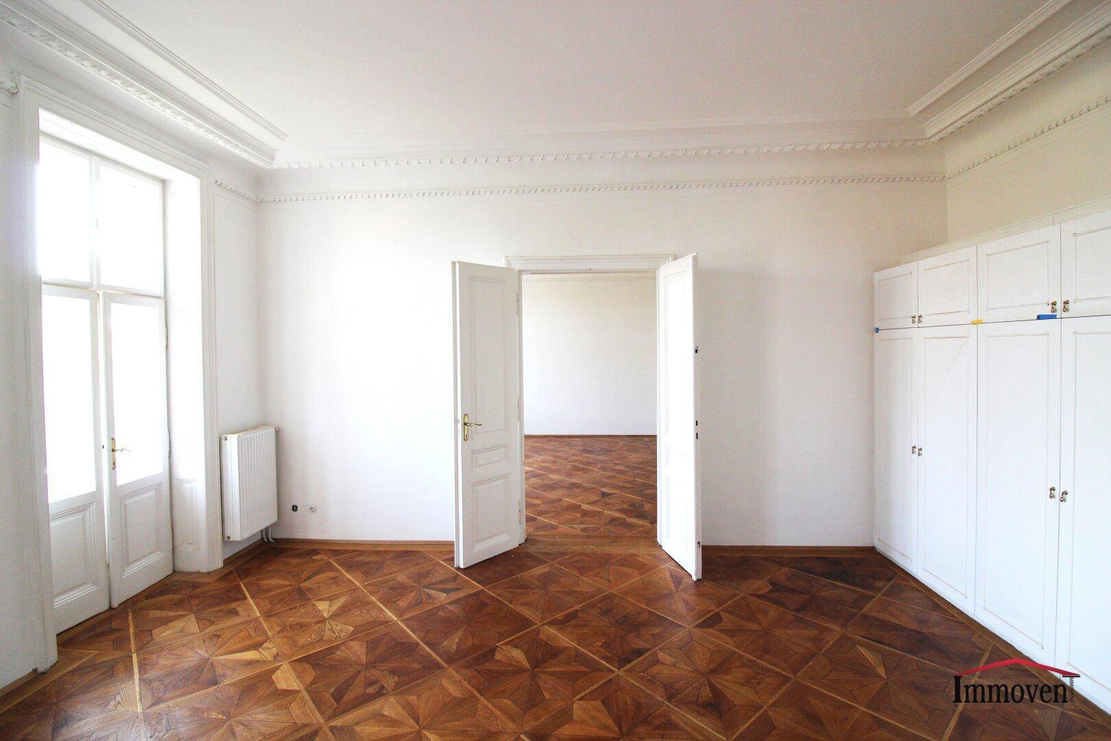 Helles Zimmer mit Parkettboden, hohen Fenstern und einer Wand mit integrierten weißen Einbauschränken.