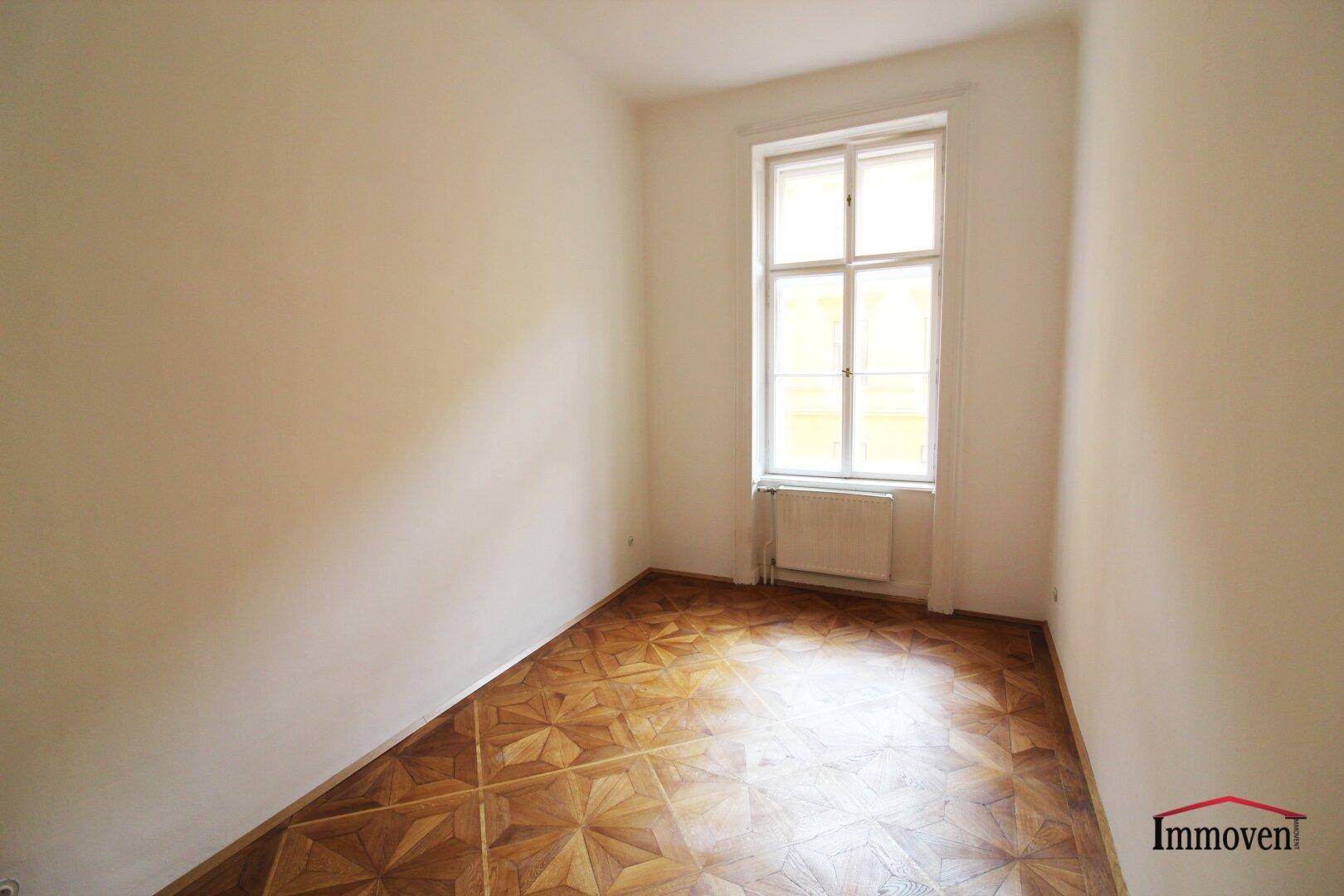 Helles, leeres Zimmer mit Parkettboden und einem hohen Fenster, das viel Tageslicht hereinlässt.