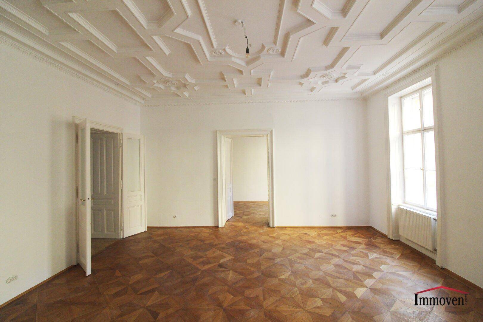 Helles Zimmer mit kunstvollem Deckenstuck, Parkettboden und hohen Fenstern. Eine Doppeltür führt in einen weiteren Raum.