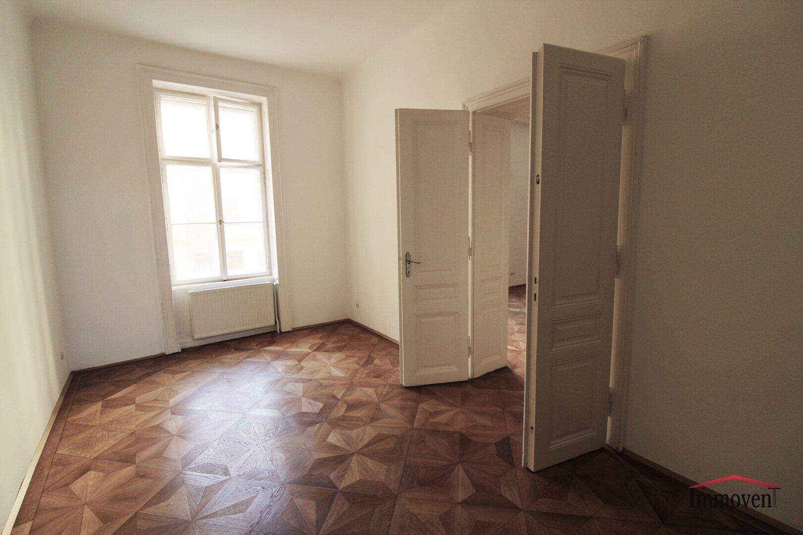 Kleineres, helles Zimmer mit Parkettboden und einem hohen Fenster. Eine Doppeltür führt in einen angrenzenden Raum.