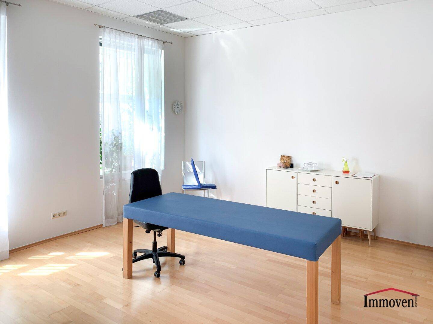 Heller Behandlungsraum oder Büro mit einem Untersuchungstisch, Bürostuhl und einem weißen Sideboard.