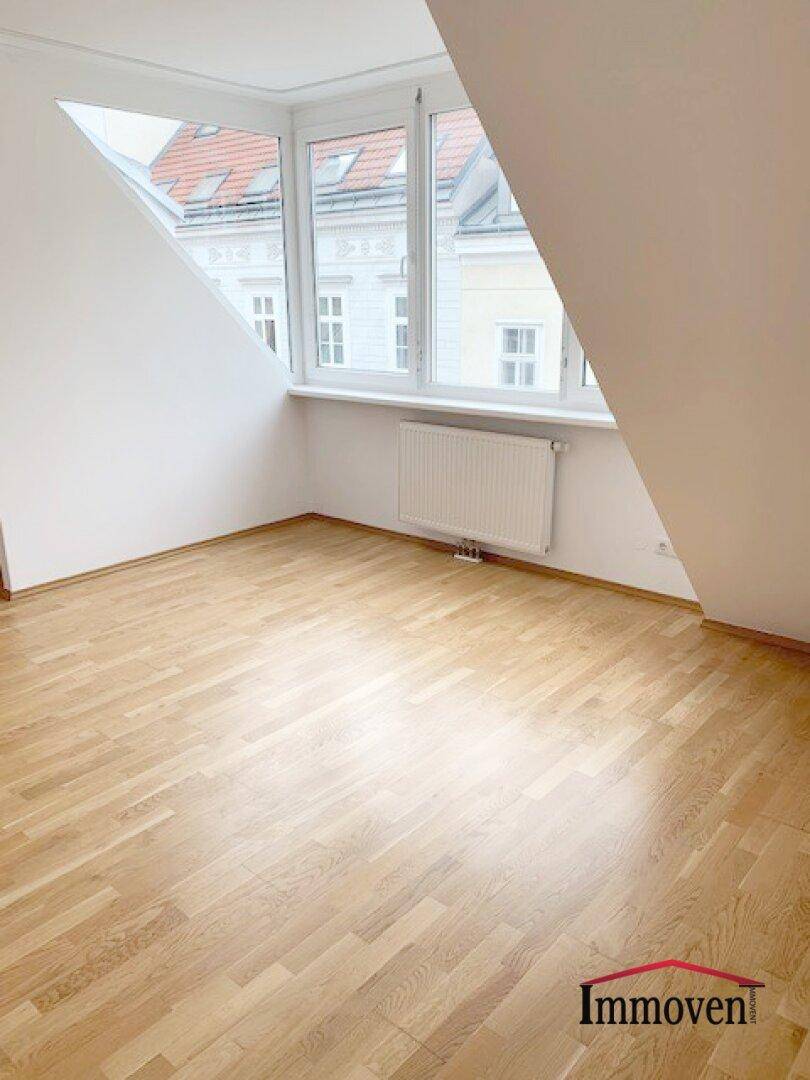 Dachgeschosszimmer mit schrägen Wänden, Parkettboden und großen Fenstern mit Blick auf die Dächer.