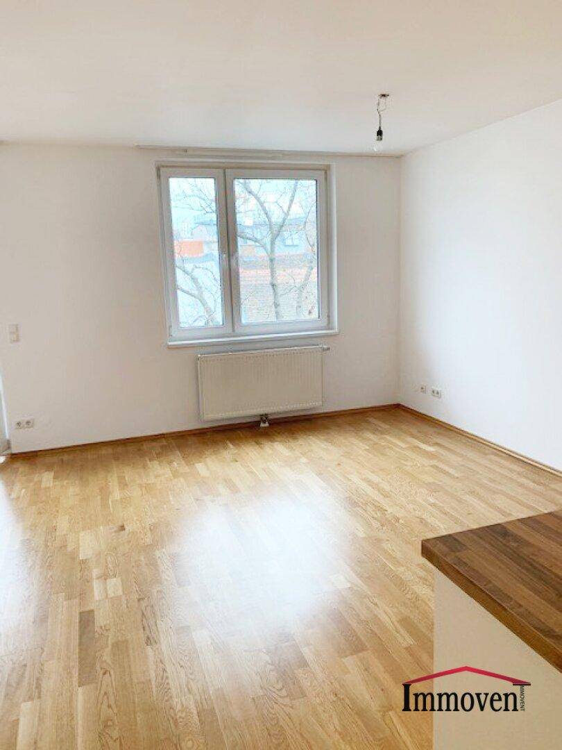 Heller, leerer Raum mit Parkettboden, weißen Wänden und einem großen Fenster mit Blick ins Grüne.