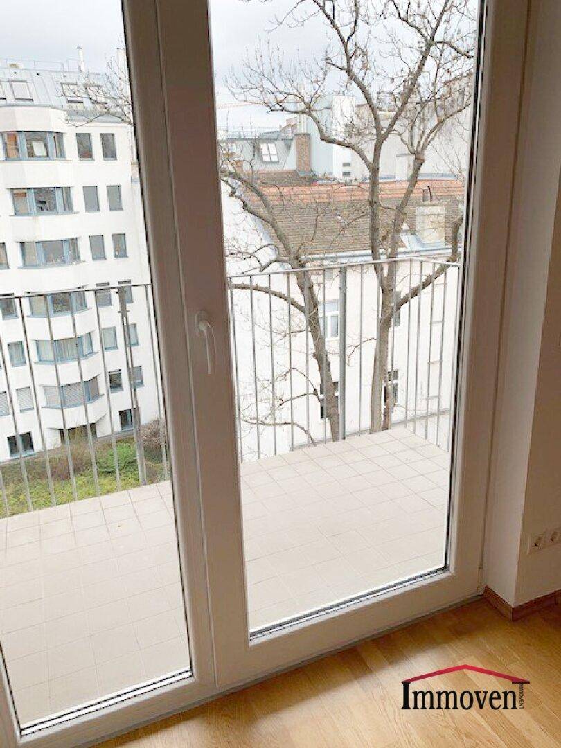 Blick vom Innenraum auf einen Balkon mit Fliesenboden und Metallgeländer, umgeben von Stadtgebäuden.