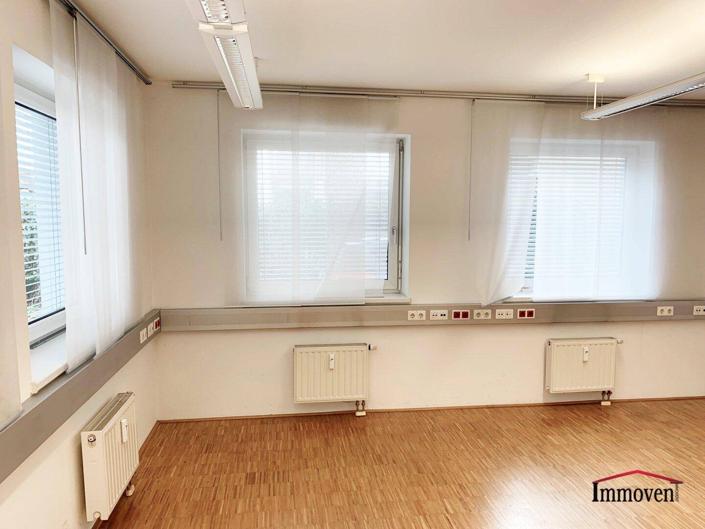 Heller Büroraum mit Parkettboden, großen Fenstern und Jalousien, bietet viel Tageslicht.