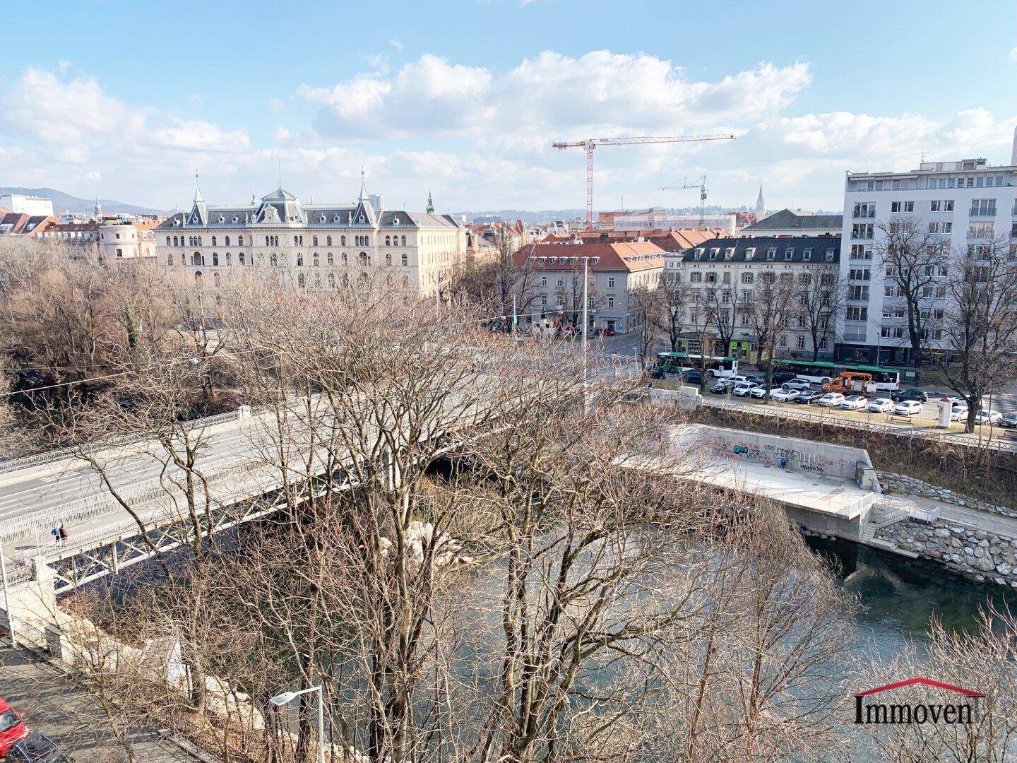 Panoramablick auf eine Brücke, den Fluss und die umliegende Stadtarchitektur unter blauem Himmel.