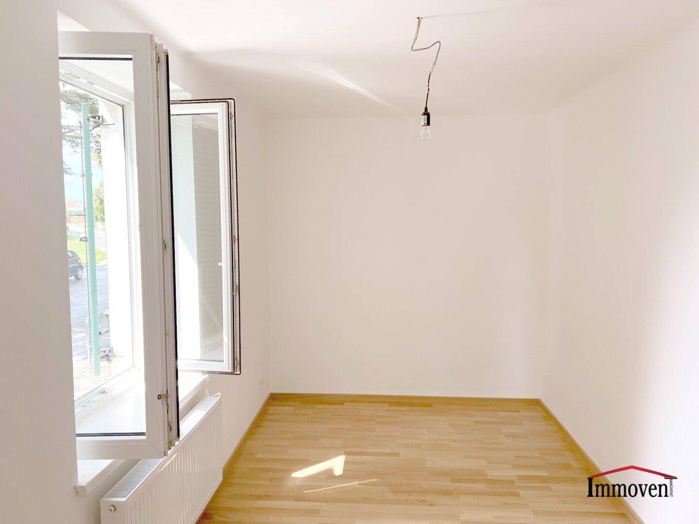 Leeres Zimmer mit hellem Holzfußboden und großen Fenstern, die viel Tageslicht hereinlassen.