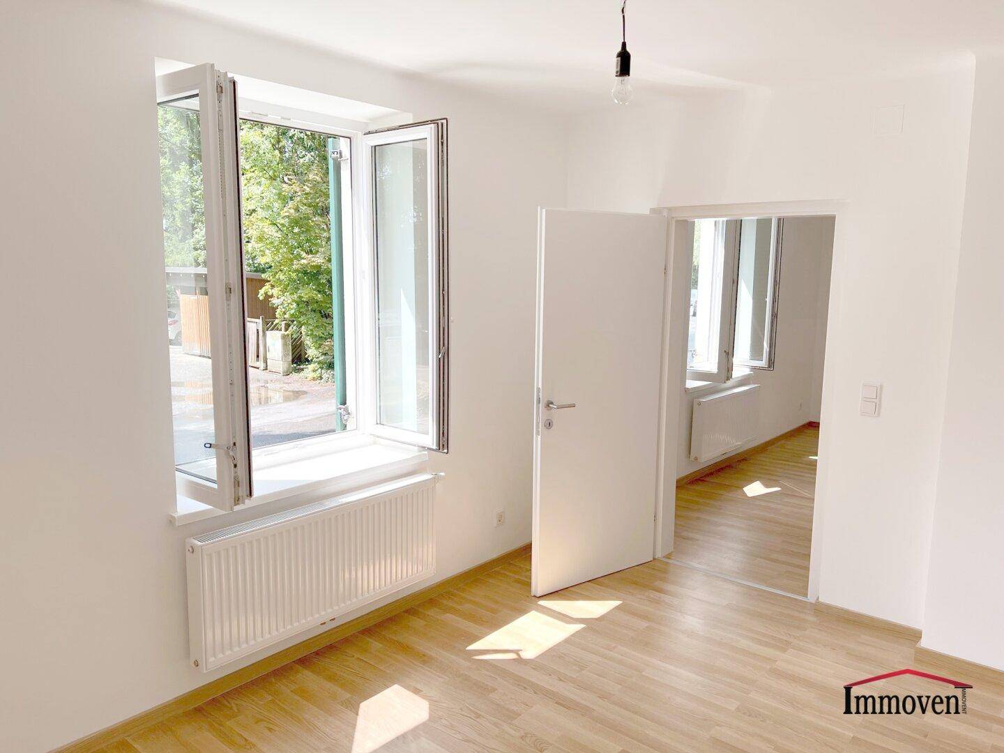 Heller Raum mit Holzfußboden, großem Fenster und offenem Zugang zu einem weiteren Zimmer.