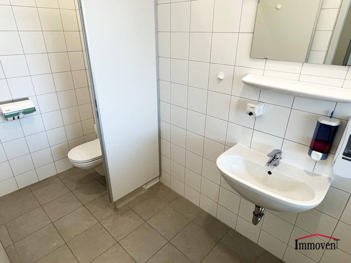 Saubere Toilettenanlage mit weißen Fliesen, Waschbecken und Trennwänden.