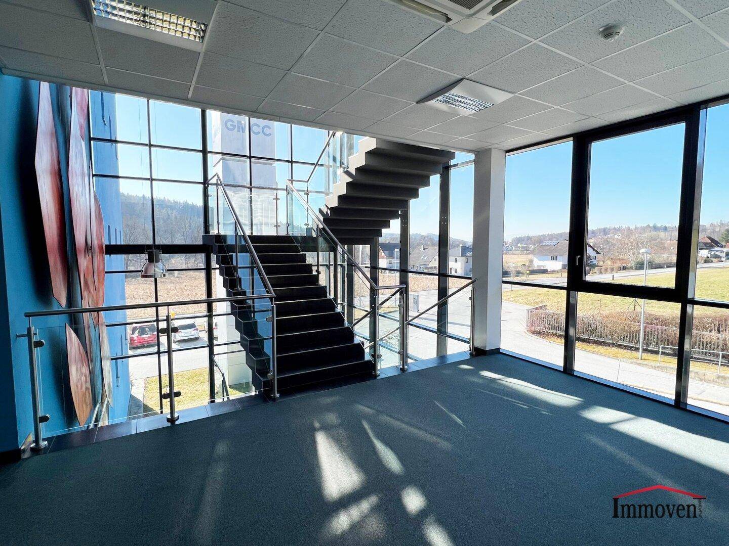Moderner Treppenaufgang mit Glasgeländer und großen Fenstern, die viel Licht hereinlassen.